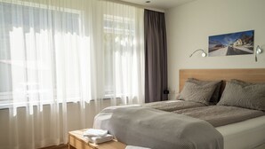 Een hotelkamer met een bed, een bankje met handdoeken, een schilderij aan de muur en dunne gordijnen.