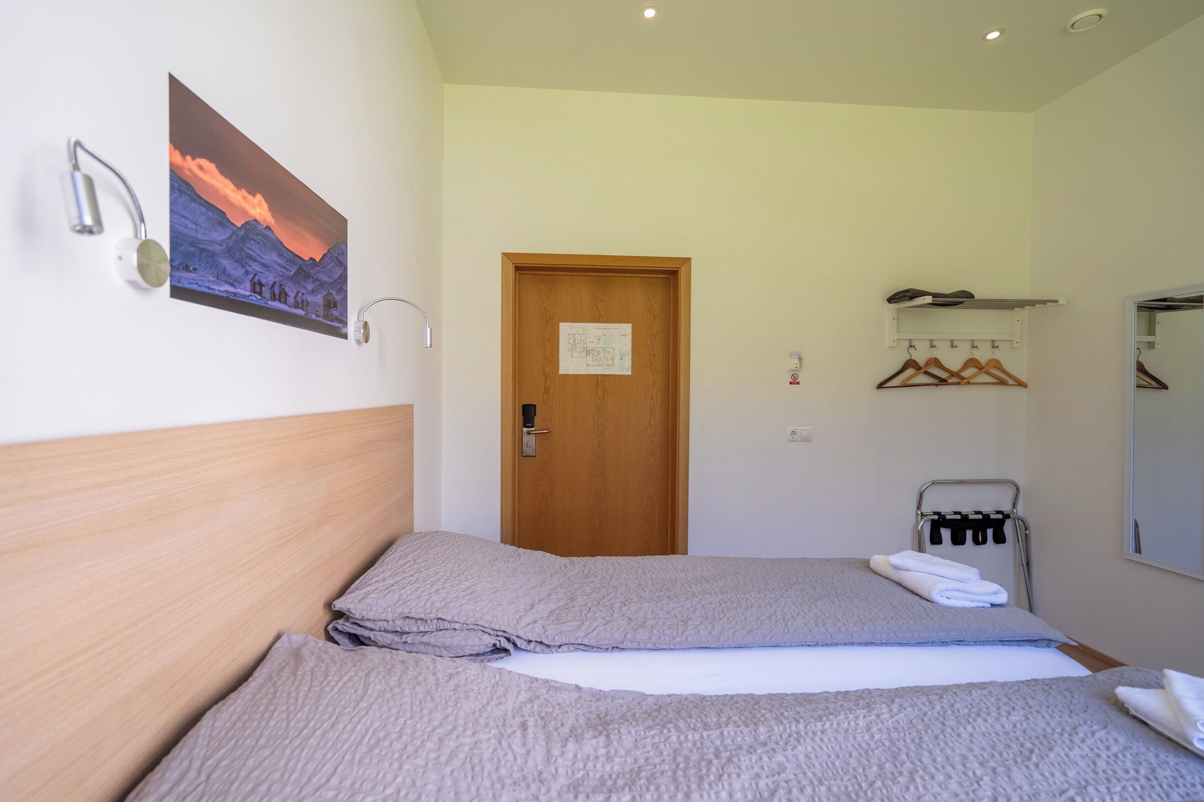 A bedroom with a wooden headboard, a bed with grey bedding, a painting of a mountain landscape, a wooden door, a coat rack with hangers, and a towel rack.