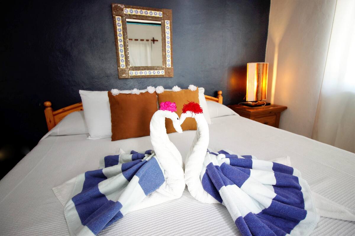 Ein ordentlich gemachtes Bett mit einem Handtuch, das zu zwei Schwänen geformt ist, ein hölzernes Kopfteil, ein Nachttisch mit Lampe und ein Spiegel an der Wand.
