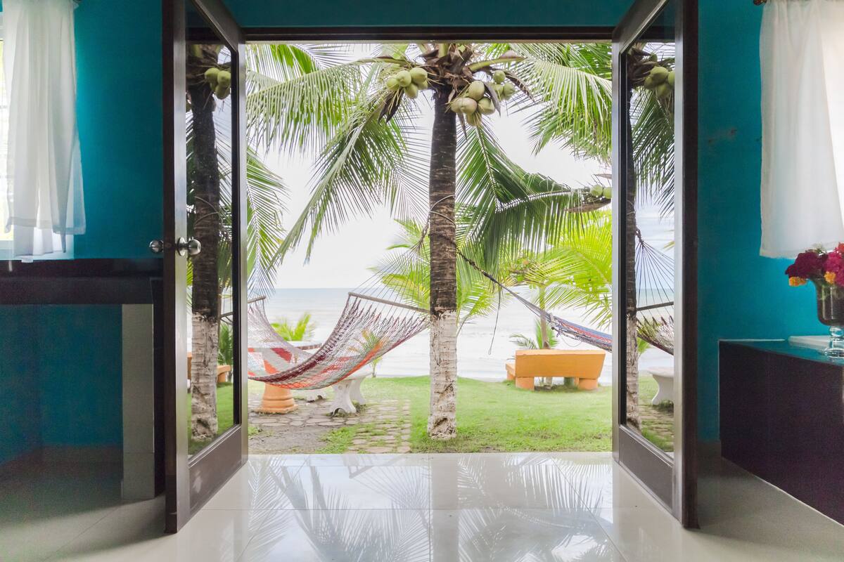 A view from a room to a beach with a hammock, palm trees, and a bench.