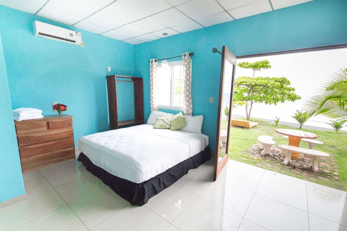 A bedroom with a bed, a dresser, a mirror, and a view of a garden with a table and chairs.