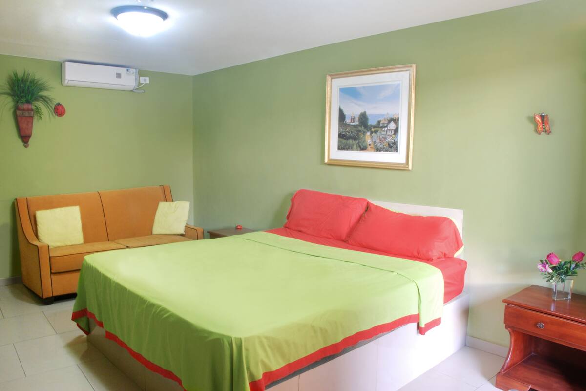 A room with a bed, a sofa, a nightstand, a potted plant, and a framed picture on the wall.