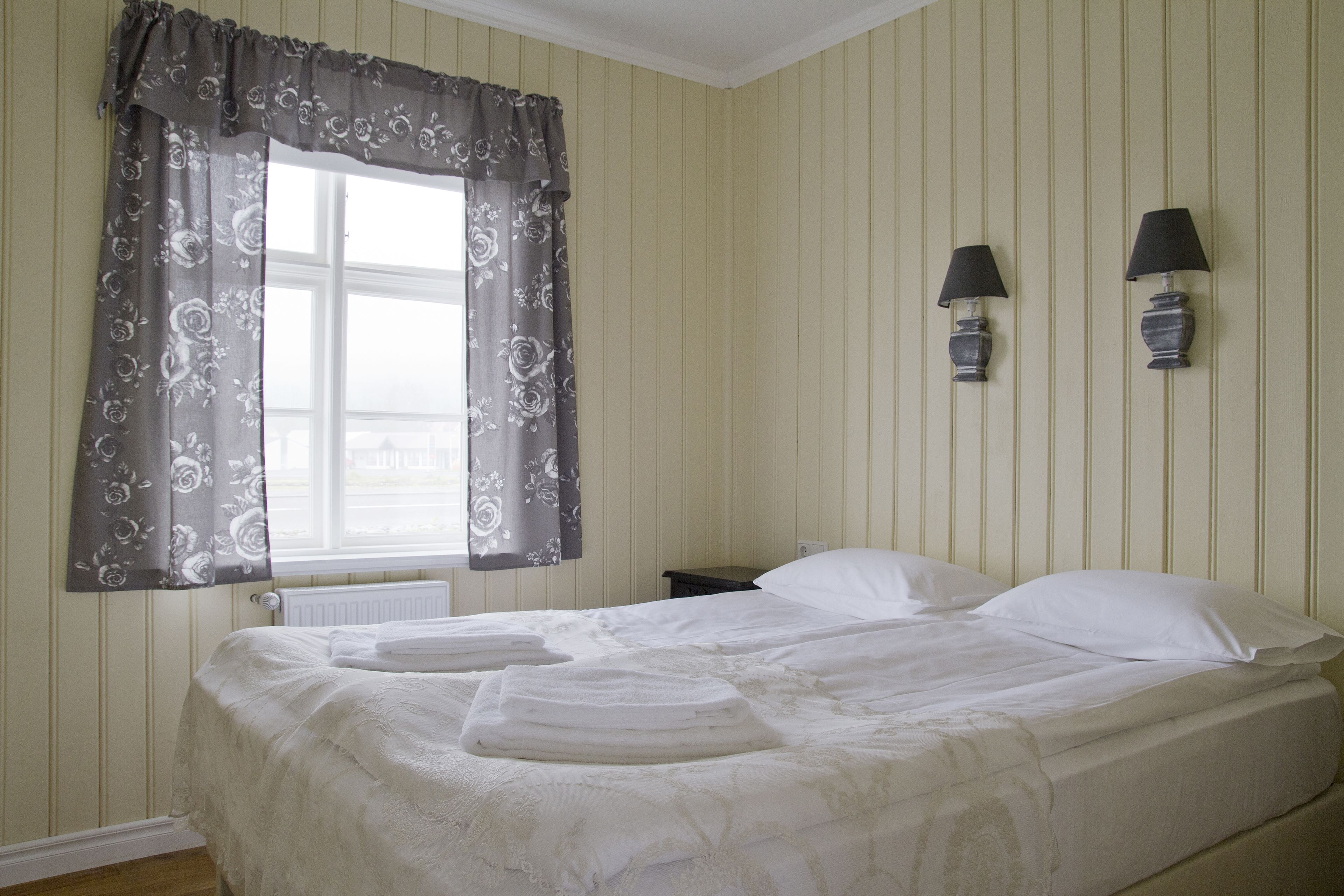 Cama bem arrumada com roupa de cama branca e cortina estampada com flores. Há duas luminárias fixadas na parede, uma de cada lado da cama.