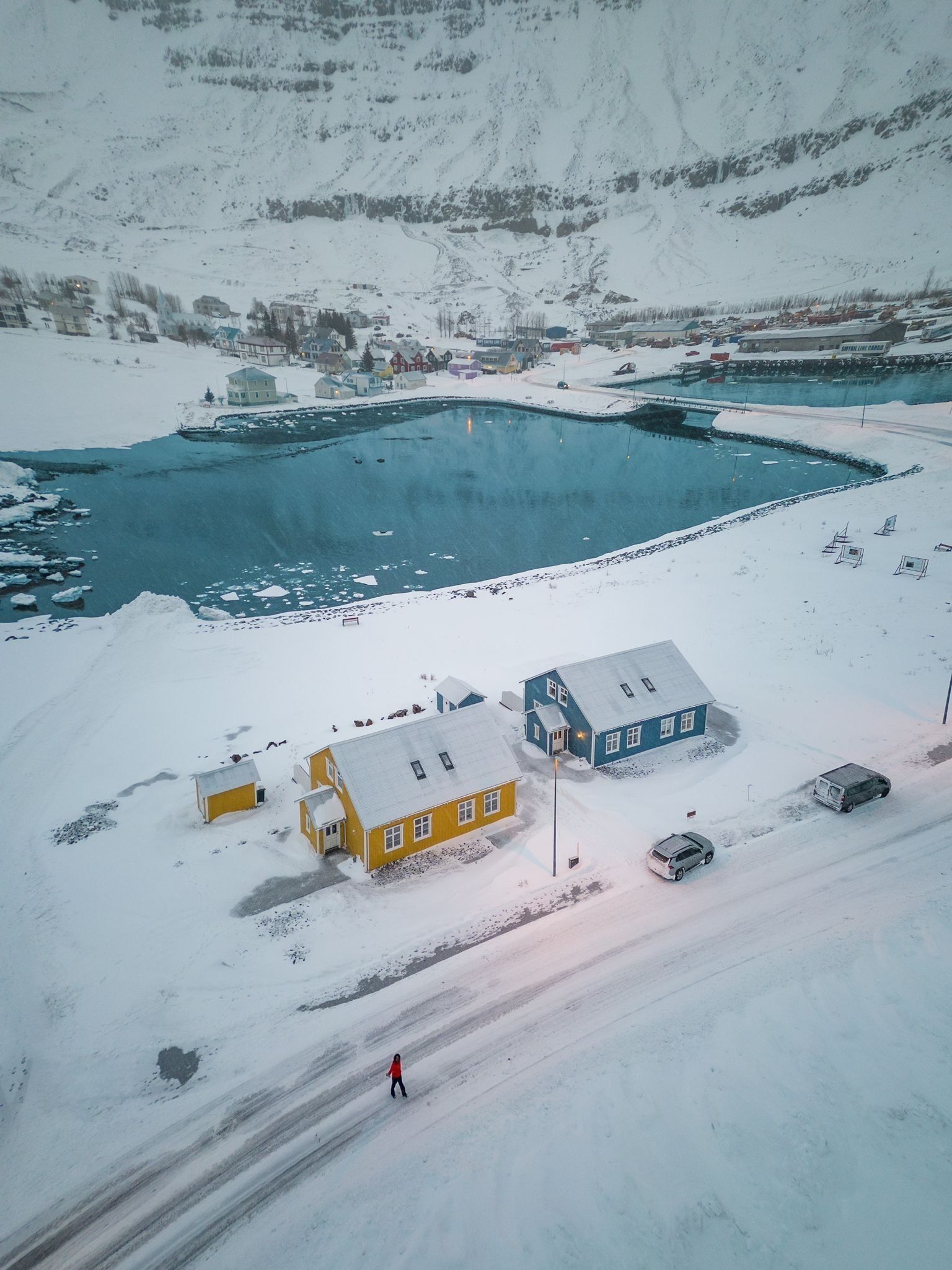 An aerial view of a snow-covered area with buildings, a frozen lake, and a person walking.