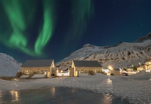 A snow-covered building with a reflective water surface in front, set against a backdrop of snow-covered mountains and the Northern Lights.