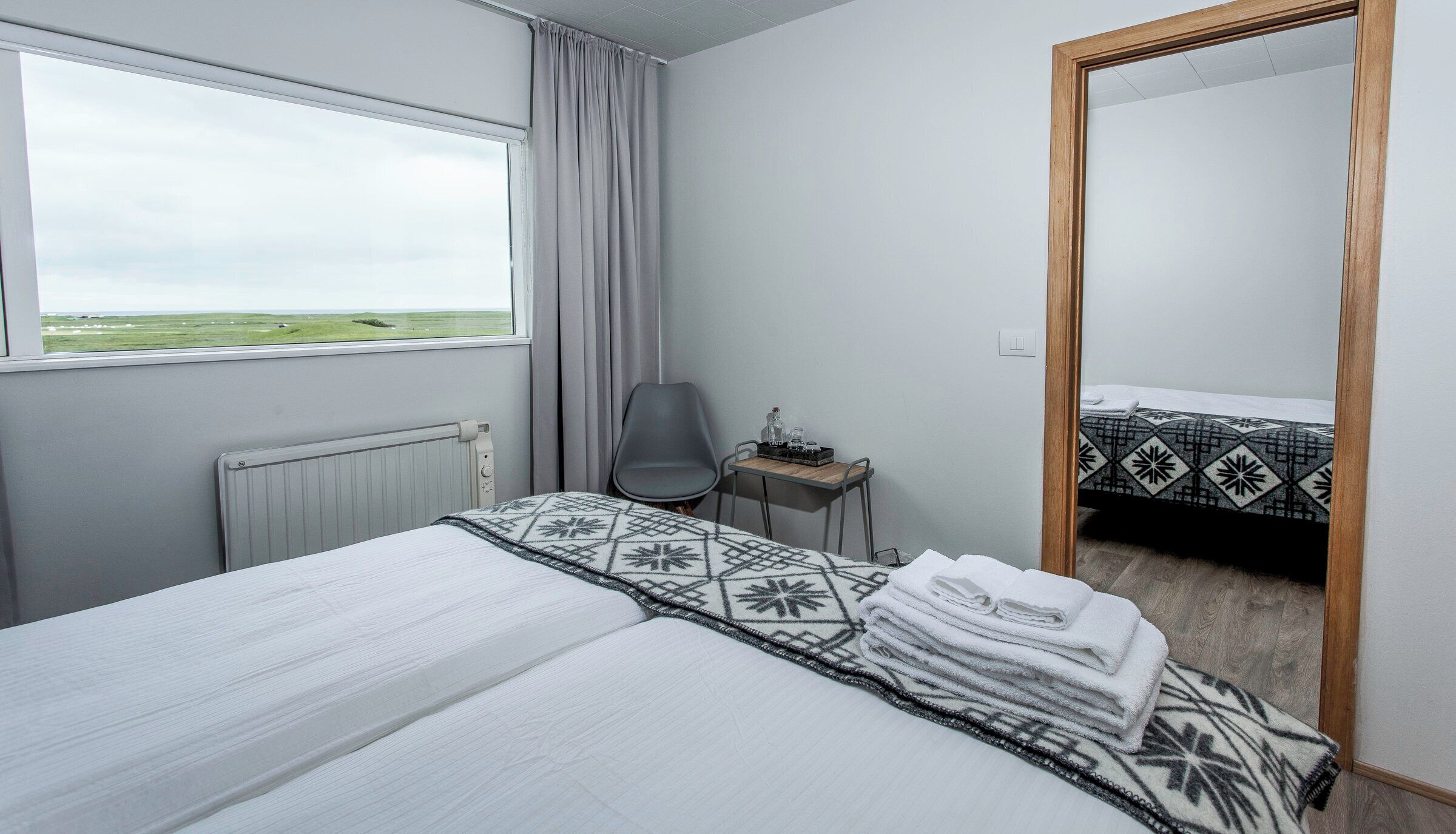 Habitación de hotel con cama, una silla, una mesita con artículos de aseo, ventana con vistas al campo y un espejo que refleja la cama.