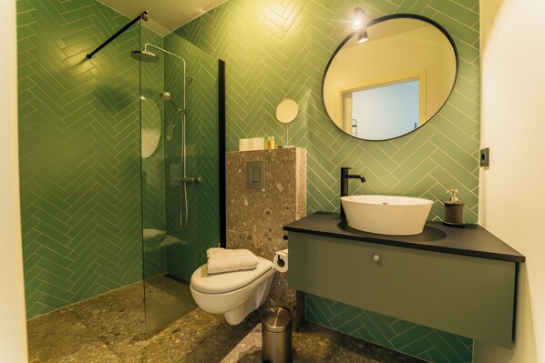 Un baño con pared de herradura verde, cabina de ducha de vidrio, inodoro, lavamanos con encimera de mármol y espejo redondo.