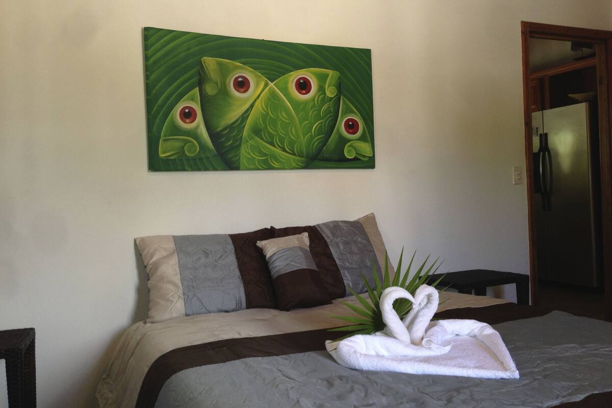 A bedroom with a bed, pillows, a painting of frogs, and a towel shaped like a swan on the bed.