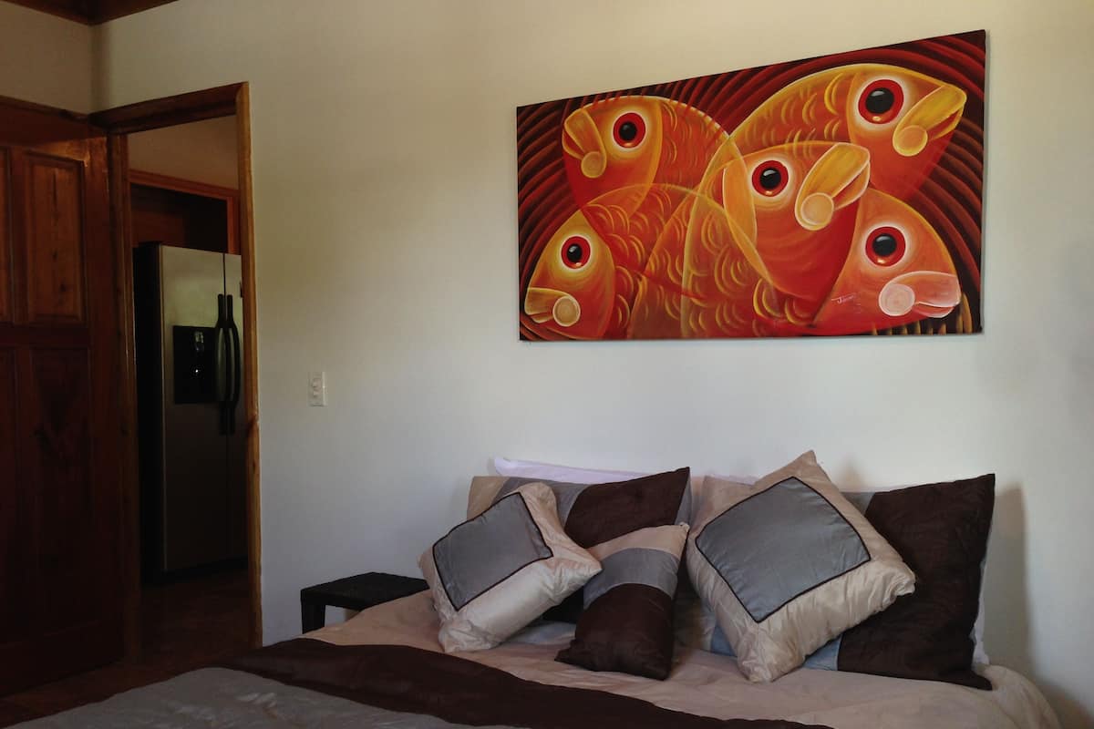 A bedroom with a bed, pillows, a painting, and a door leading to another room.