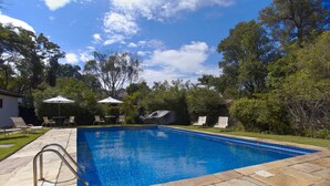 Ein rechteckiger Pool mit einer Steindeck, umgeben von Liegestühlen und Sonnenschirmen, eingebettet in einen angelegten Garten mit Bäumen und blauem Himmel.