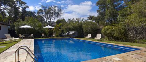 Piscina rectangular con solárium de piedra, rodeada de sillones de descanso y sombrillas, ubicada en un jardín ajardinado con árboles y un cielo despejado.