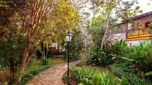 Ein steiniger Pfad schlängelt sich durch einen üppigen Garten mit verschiedenen Pflanzen und einem traditionellen Gebäude mit einer gelb-weißen Fassade.