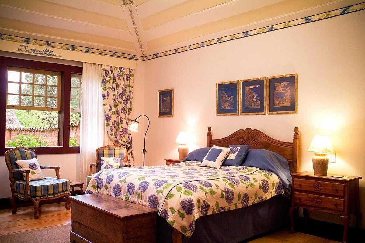 A bedroom with a wooden bed, two bedside tables with lamps, a chair, a wooden chest, and a window with curtains.