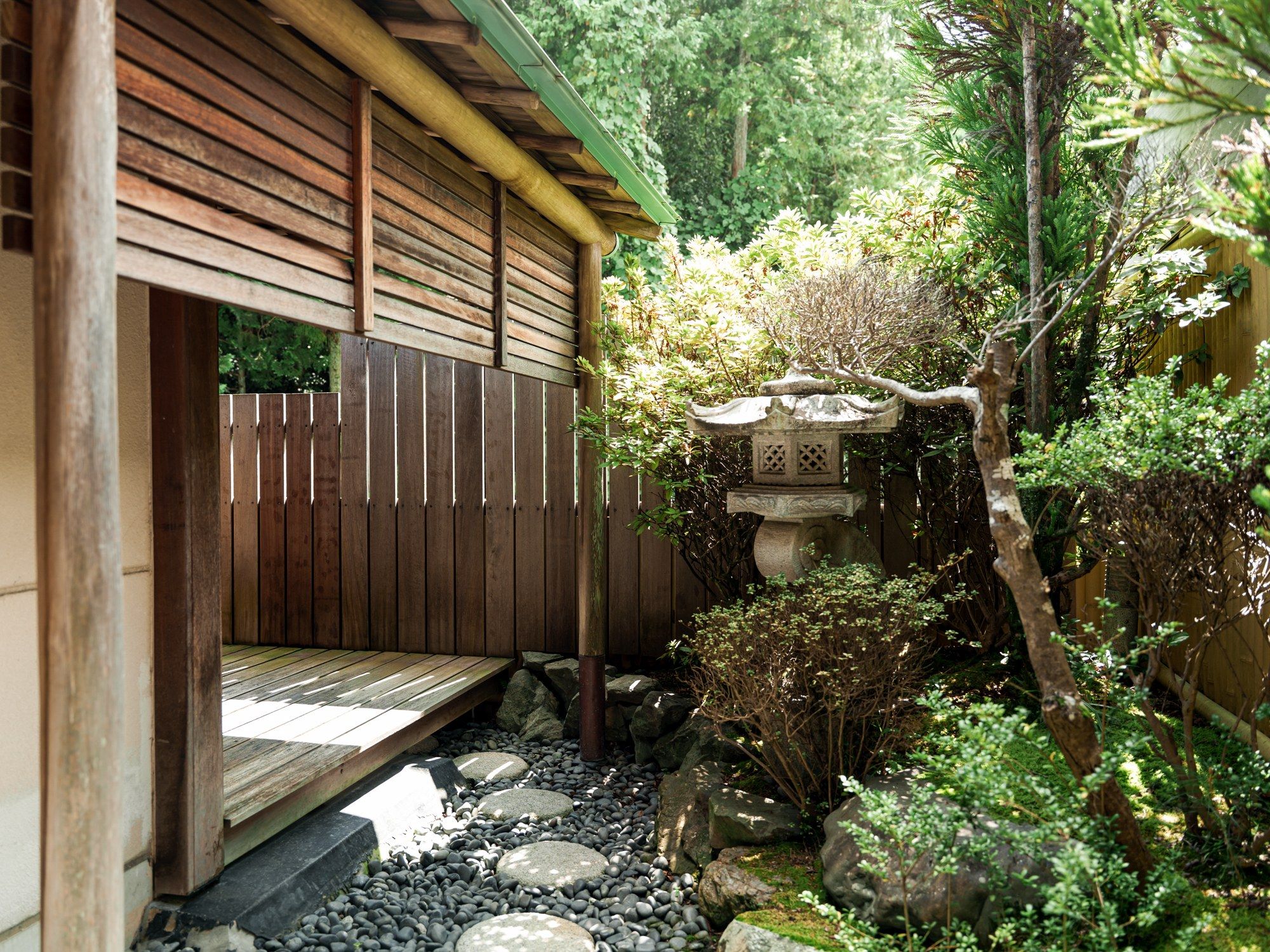 A wooden structure with a sloped roof, surrounded by a garden with rocks, plants, and a stone water feature.