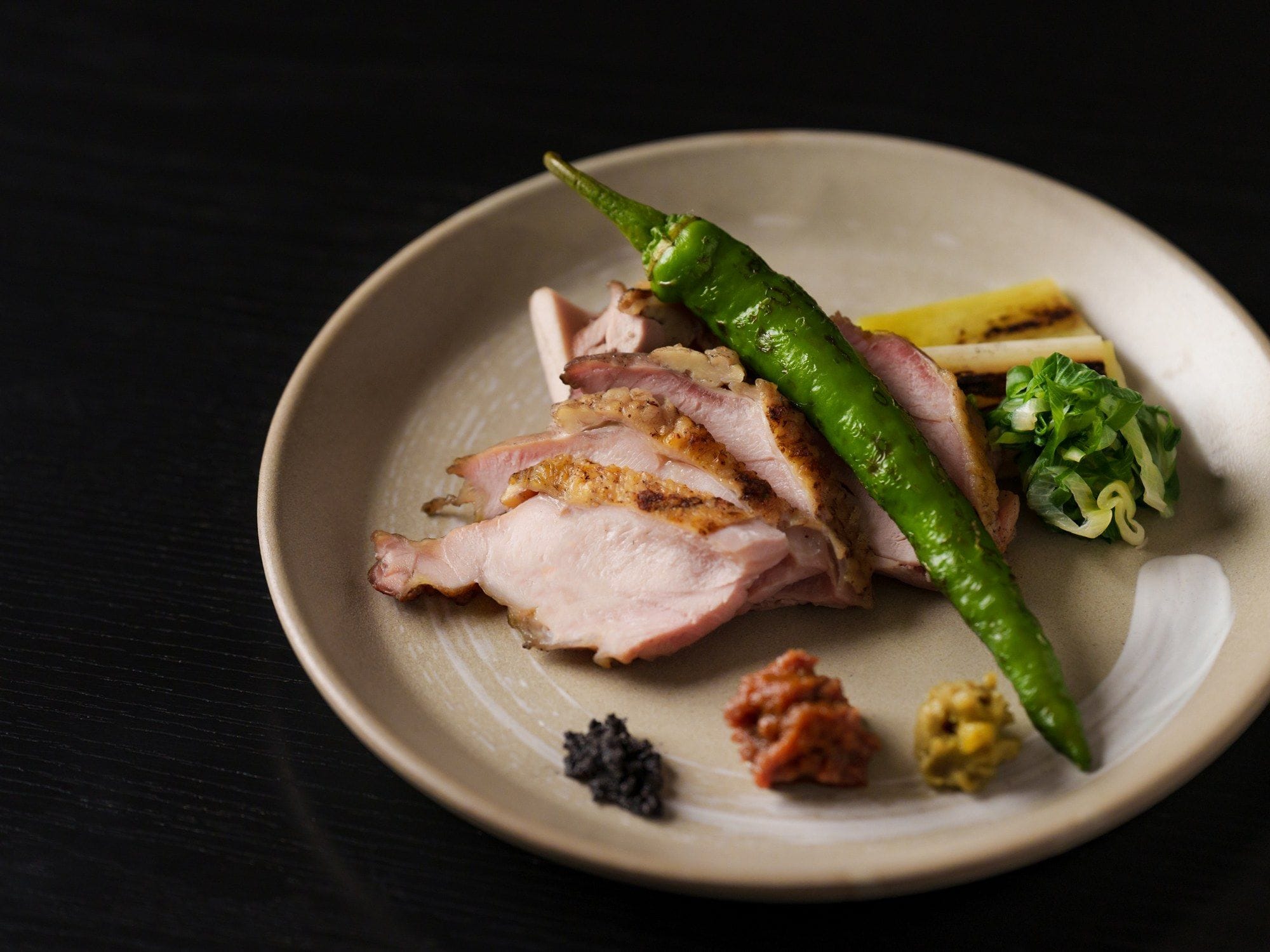Un plato con carne en rodajas, un chile verde y unas hojas verdes sobre una superficie oscura.
