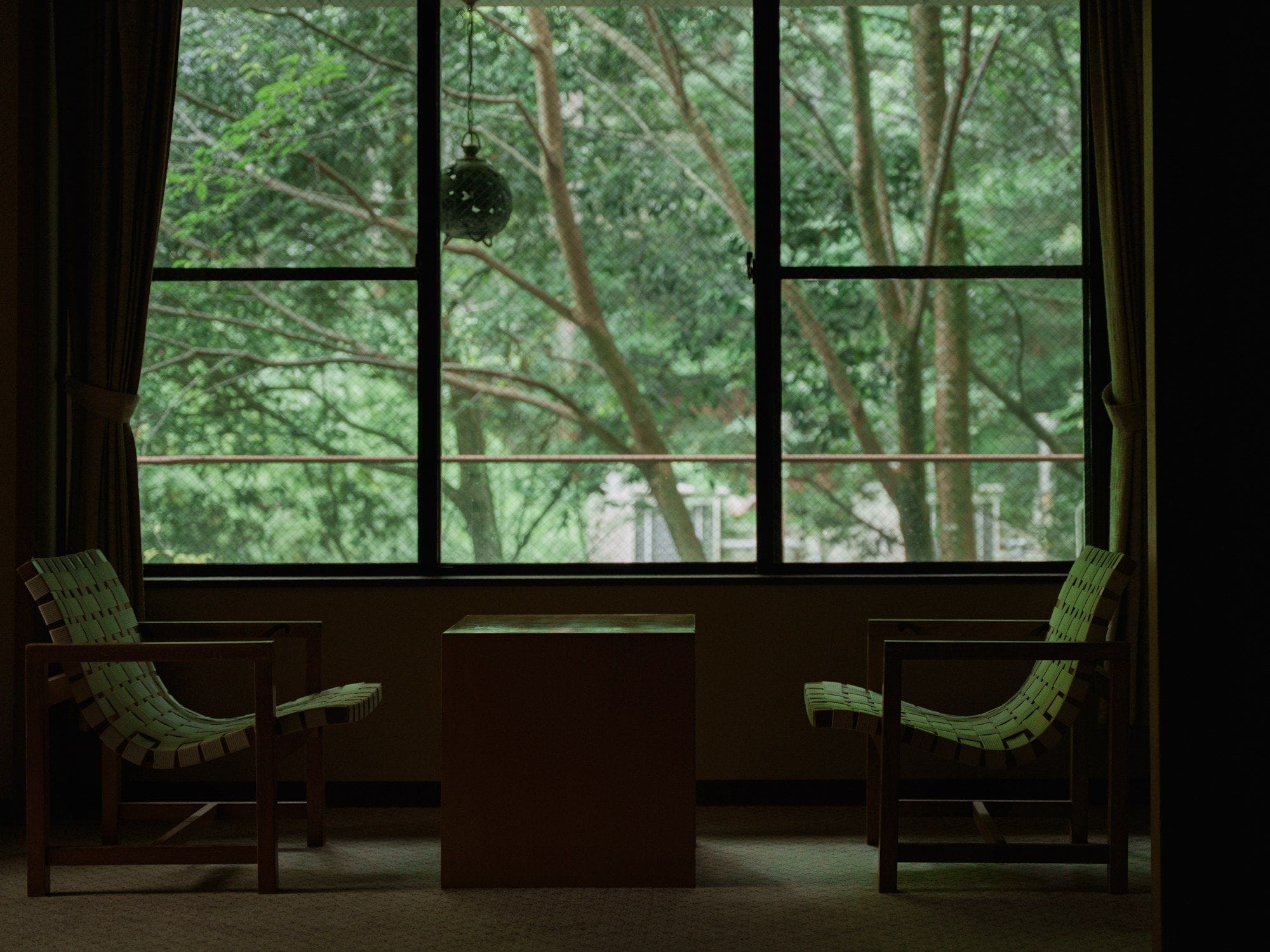 Una habitación con dos sillas y una mesa, un ventanal con vista a un bosque y una lámpara colgante.