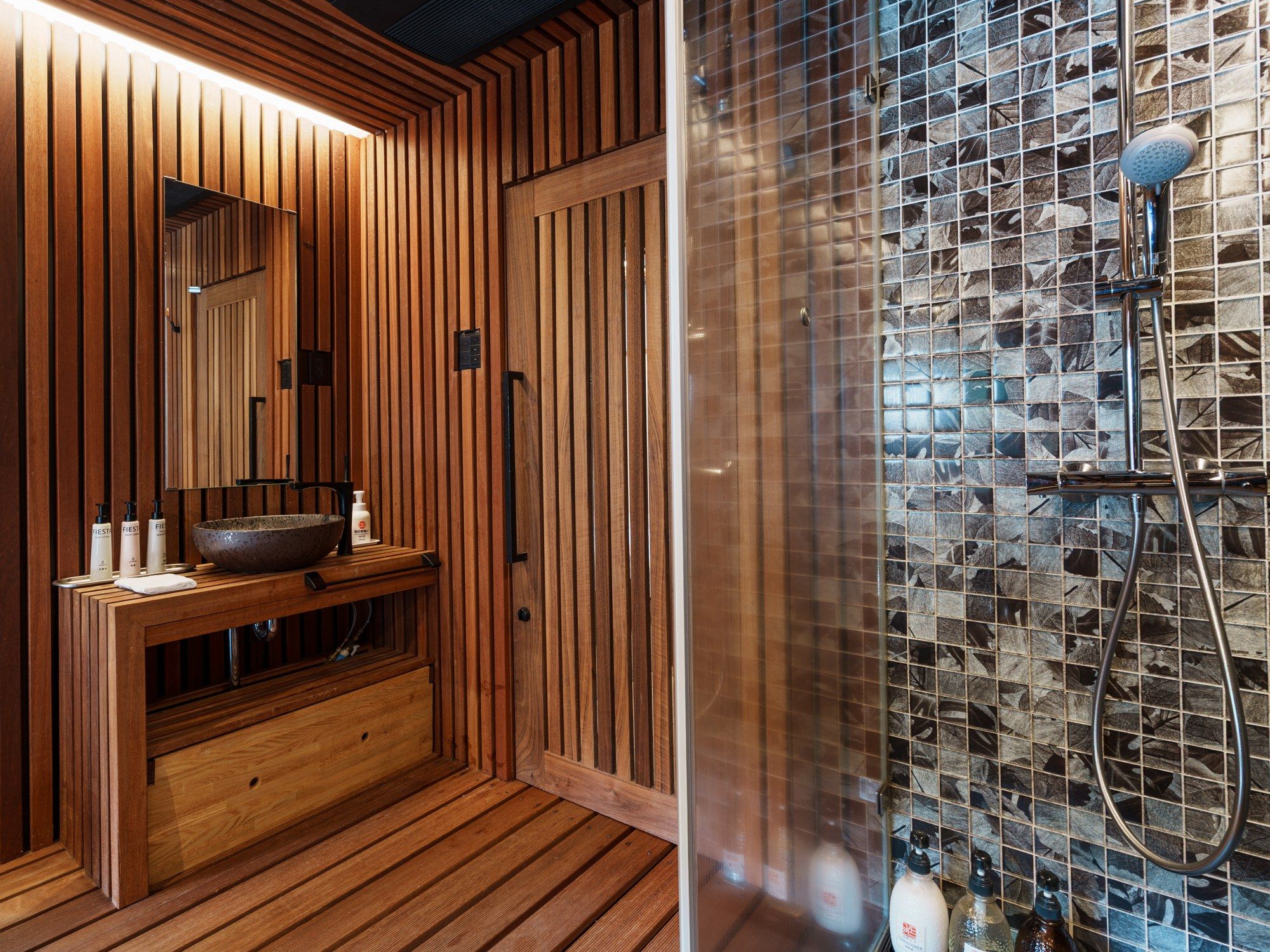 Una sauna de madera con un espejo grande, un banco de madera y un área de ducha acristalada con pared de azulejos.