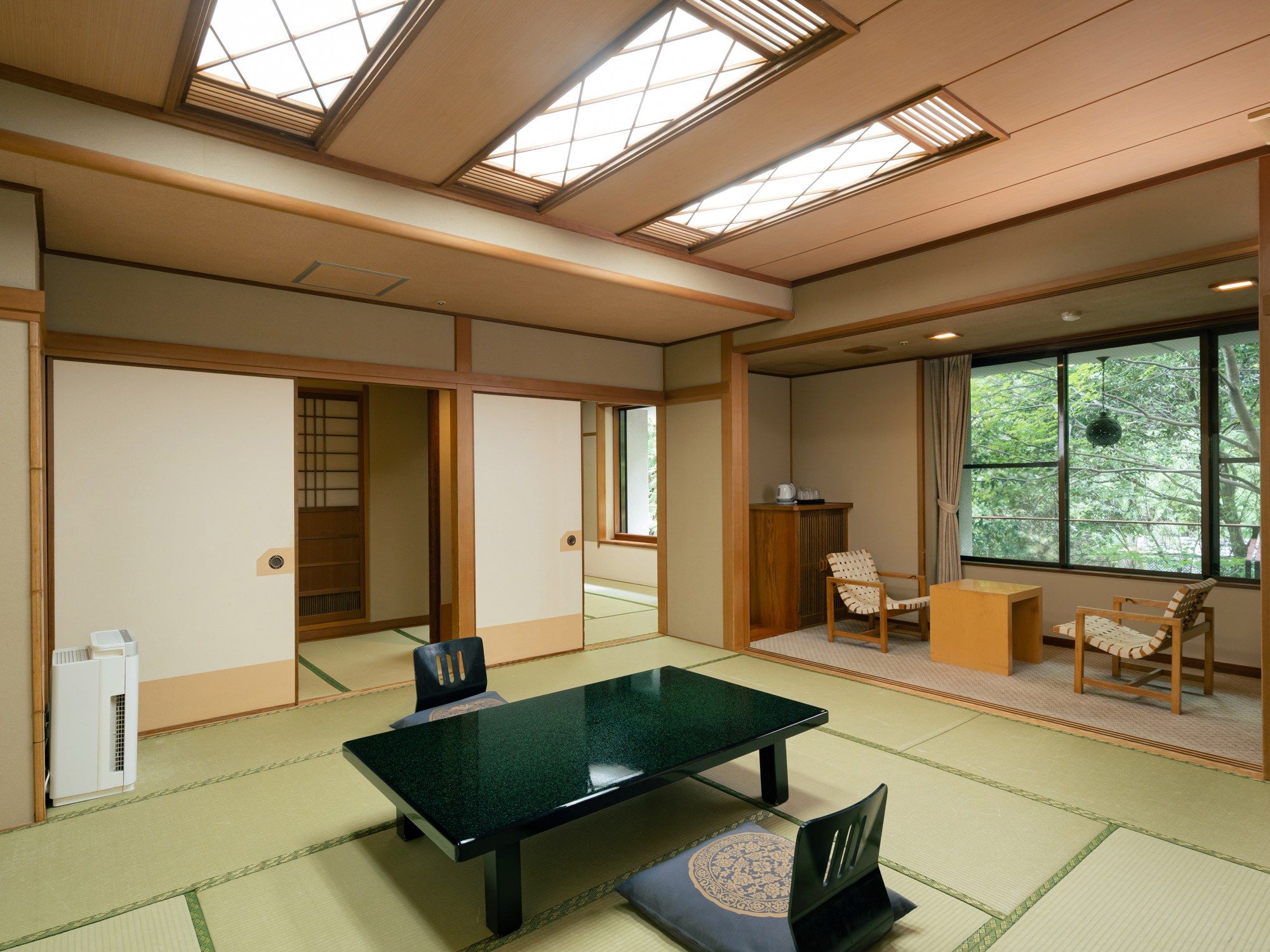 Une pièce de style japonais traditionnel, avec un sol recouvert de tatamis, une table basse, des chaises et une vue sur la verdure grâce à de grandes fenêtres.