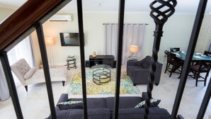 Un vaste espace de vie comprenant un canapé, un fauteuil et une table basse en verre. On aperçoit une table à manger avec des chaises au fond.