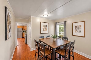 A dining area with a wooden table and chairs, framed artwork on the walls, and a view into the kitchen.