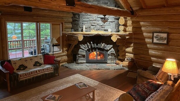 A log cabin interior with a stone fireplace, wooden furniture, and a view through large windows. - Authentic Hand-Crafted Mountain Cabin w/ Amazing Lake View ! (Somers)