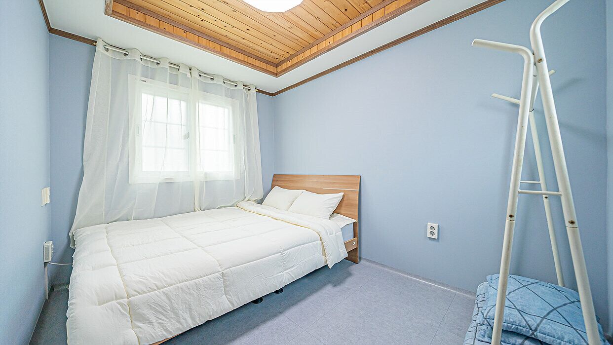 Een slaapkamer met een bed, een raam met dunne gordijnen, een houten plafond en een witte trap.