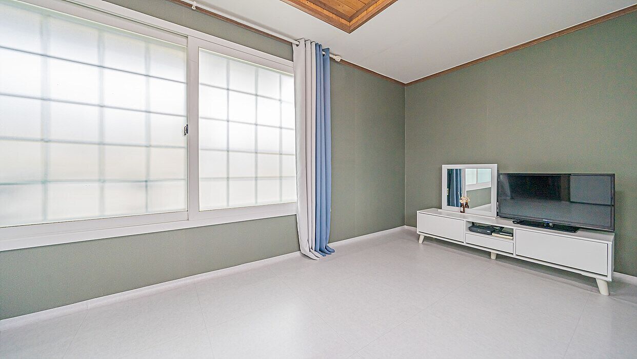 A room with a large window, a television on a white cabinet, and a mirror on the wall.