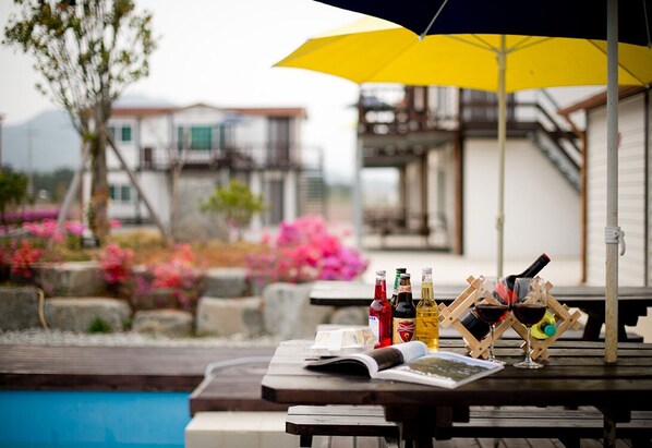 A poolside table with bottles and glasses, an umbrella, and a book. - Wando Sol Beach Pension (Wando)