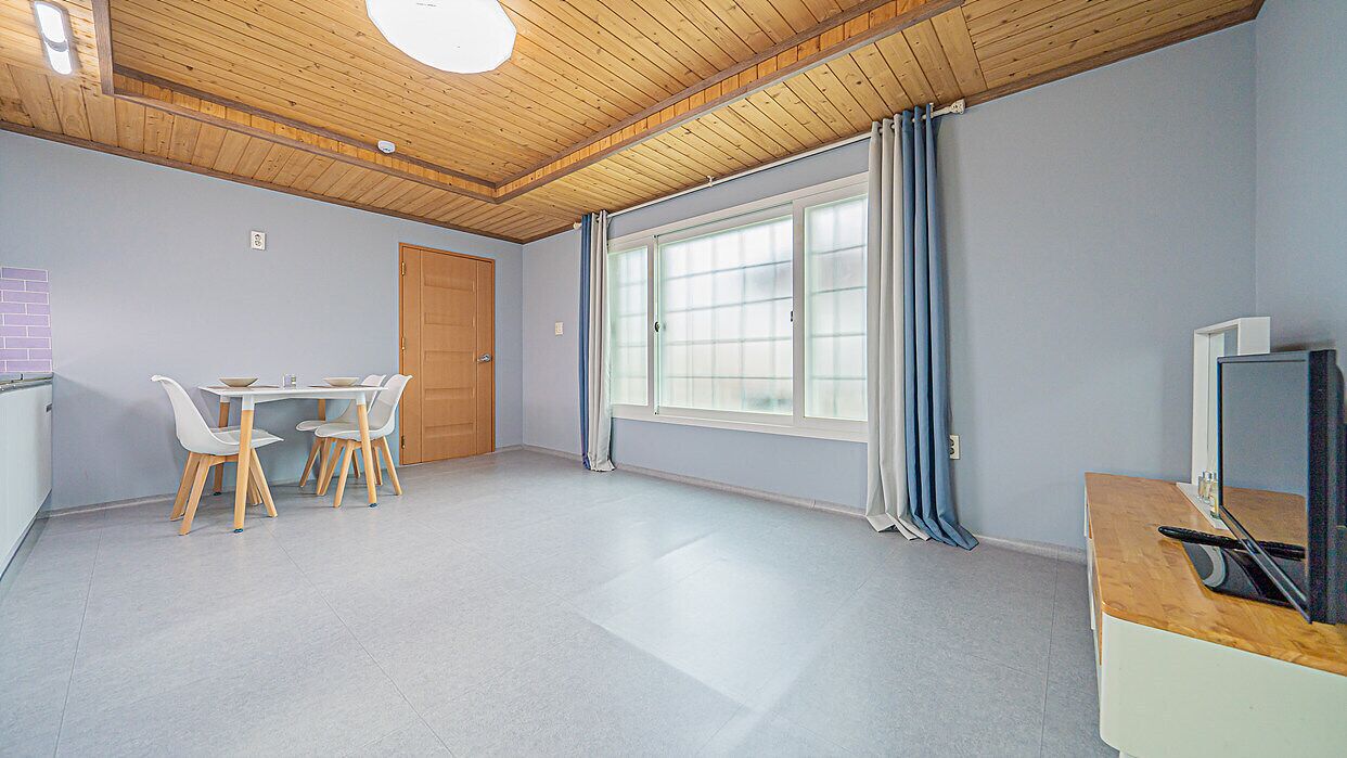 A spacious room with a wooden ceiling, a large window with curtains, a dining table with chairs, and a television on a wooden cabinet.