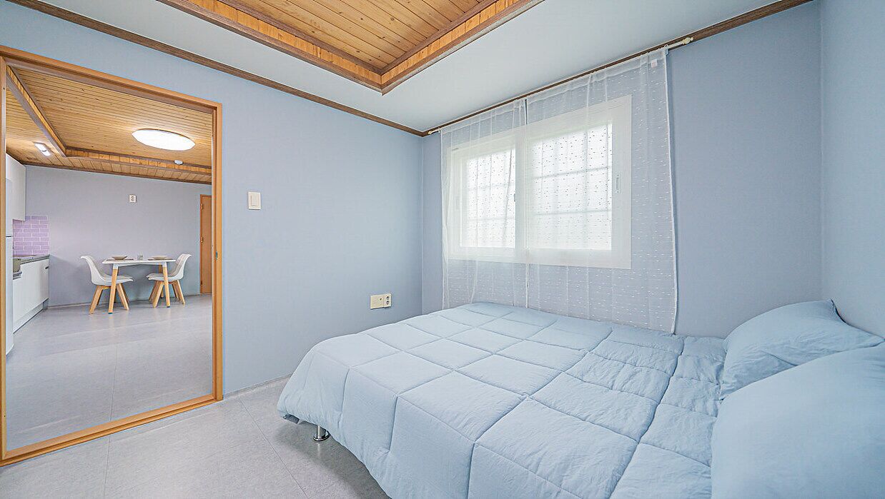 Un dormitorio con cama, una ventana con cortinas translúcidas, techo de madera y una pequeña mesa con sillas en la habitación contigua.