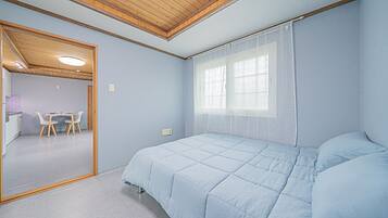 Un dormitorio con cama, una ventana con cortinas translúcidas, techo de madera y una pequeña mesa con sillas en la habitación contigua.