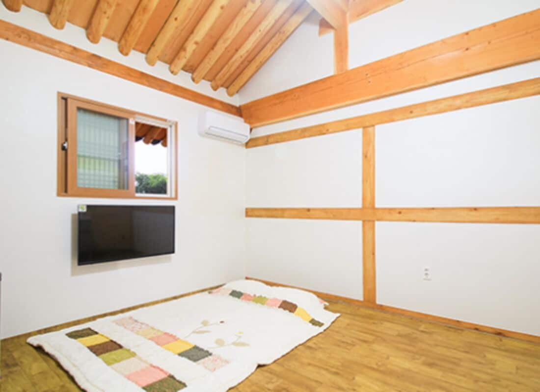 A room with wooden beams, a window, a flat-screen TV, and a quilt on the floor.