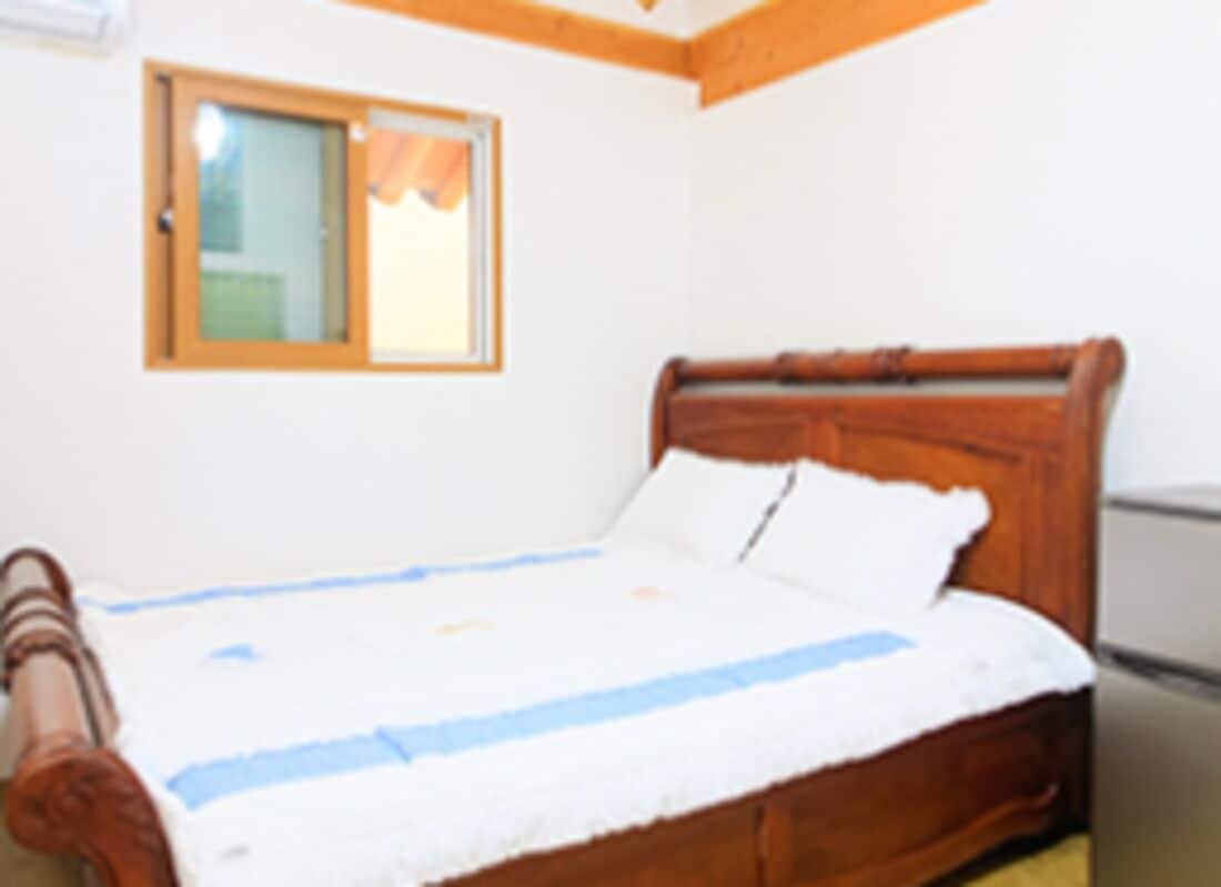Une chambre avec un lit en bois, du linge de lit blanc, une fenêtre avec un rideau et un réfrigérateur.