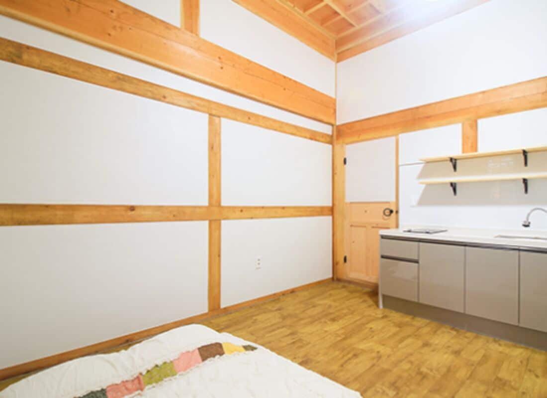 Un dormitorio con techo de madera, una cama, una pequeña kitchenette con un lavamanos y armarios, y una ventana.