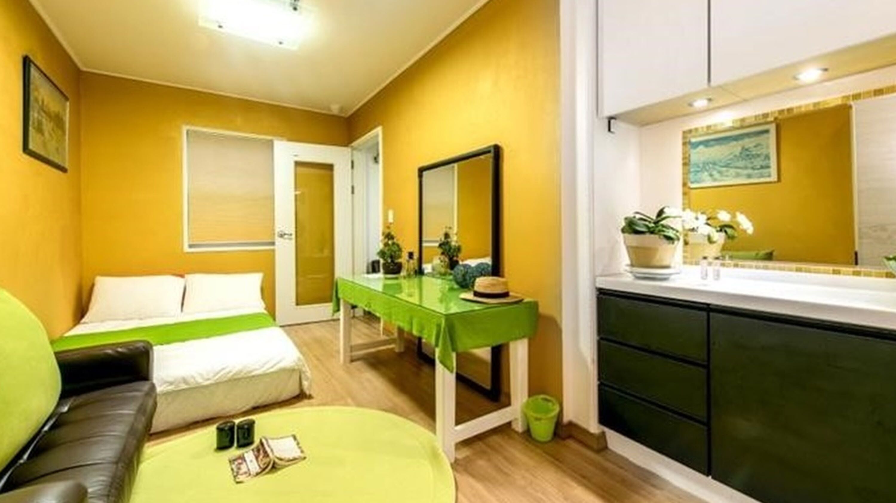 Habitación de hotel con una cama, un sofá verde, una mesita con un sombrero y una zona de cocina con un fregadero y armarios.