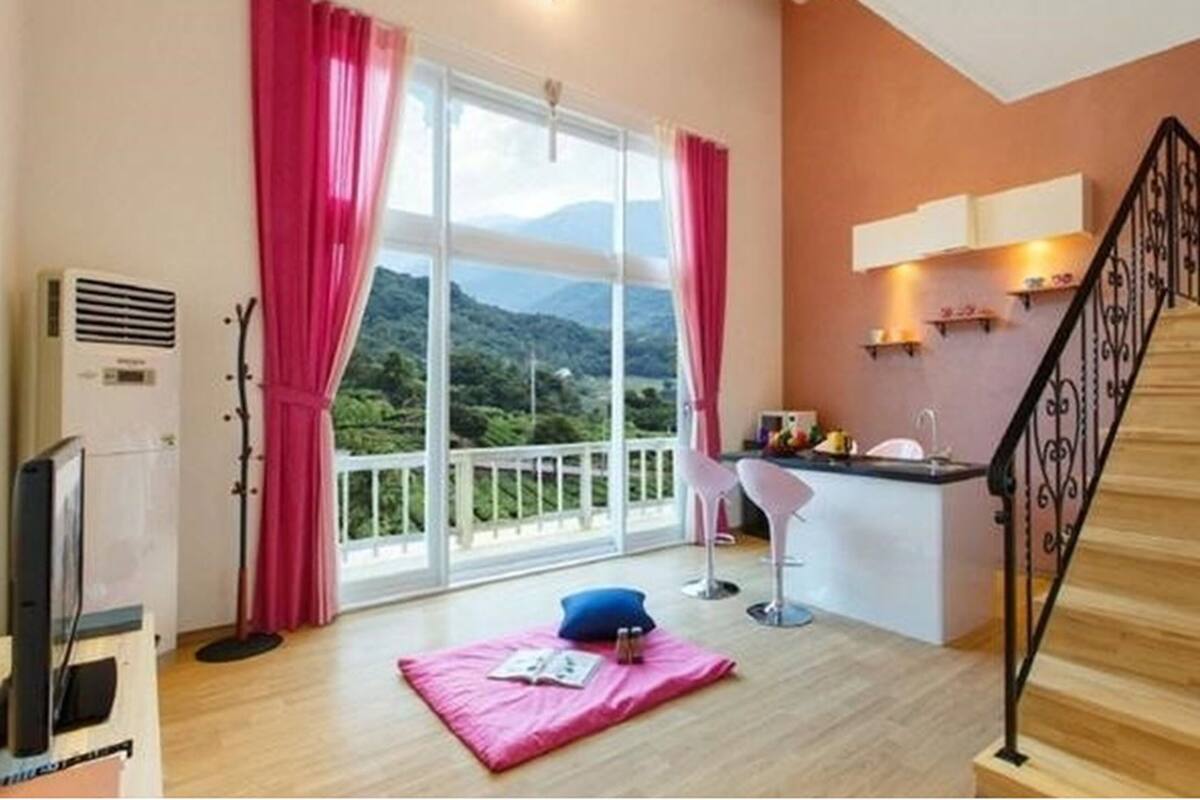 Una habitación con un amplio ventanal corredera que da a un balcón con vistas a la montaña. Hay una pequeña kitchenette con un fregadero y una silla.