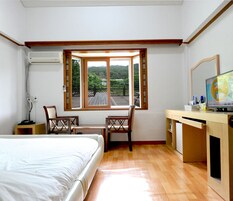 Un dormitorio con cama, escritorio y dos sillas, televisor y ventana con vista a follaje.