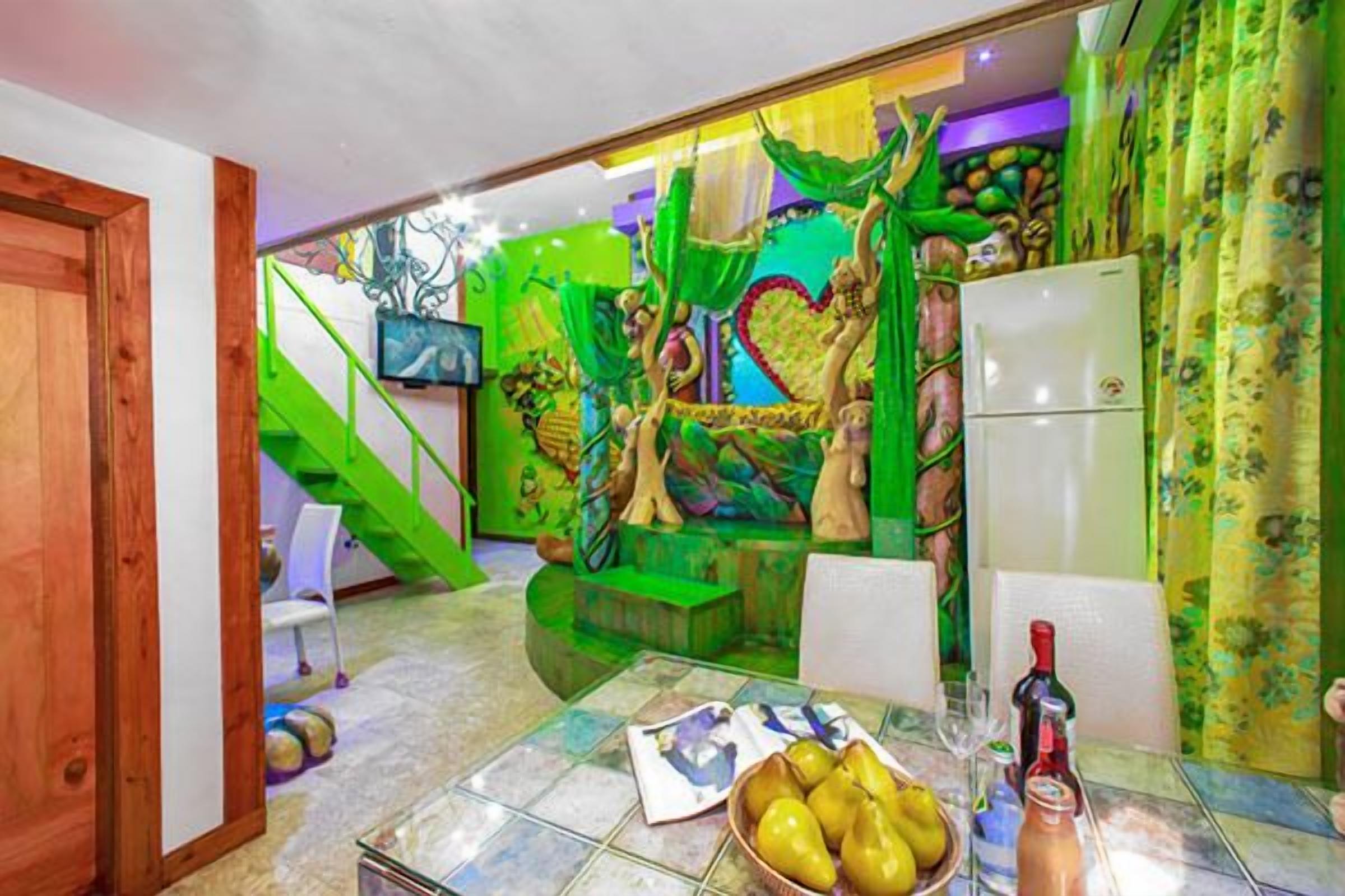 A room with a green-themed mural, a dining table with fruit, and a refrigerator.