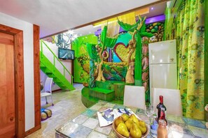 Une pièce décorée d’une fresque aux motifs verts, une table à manger avec des fruits et un réfrigérateur.