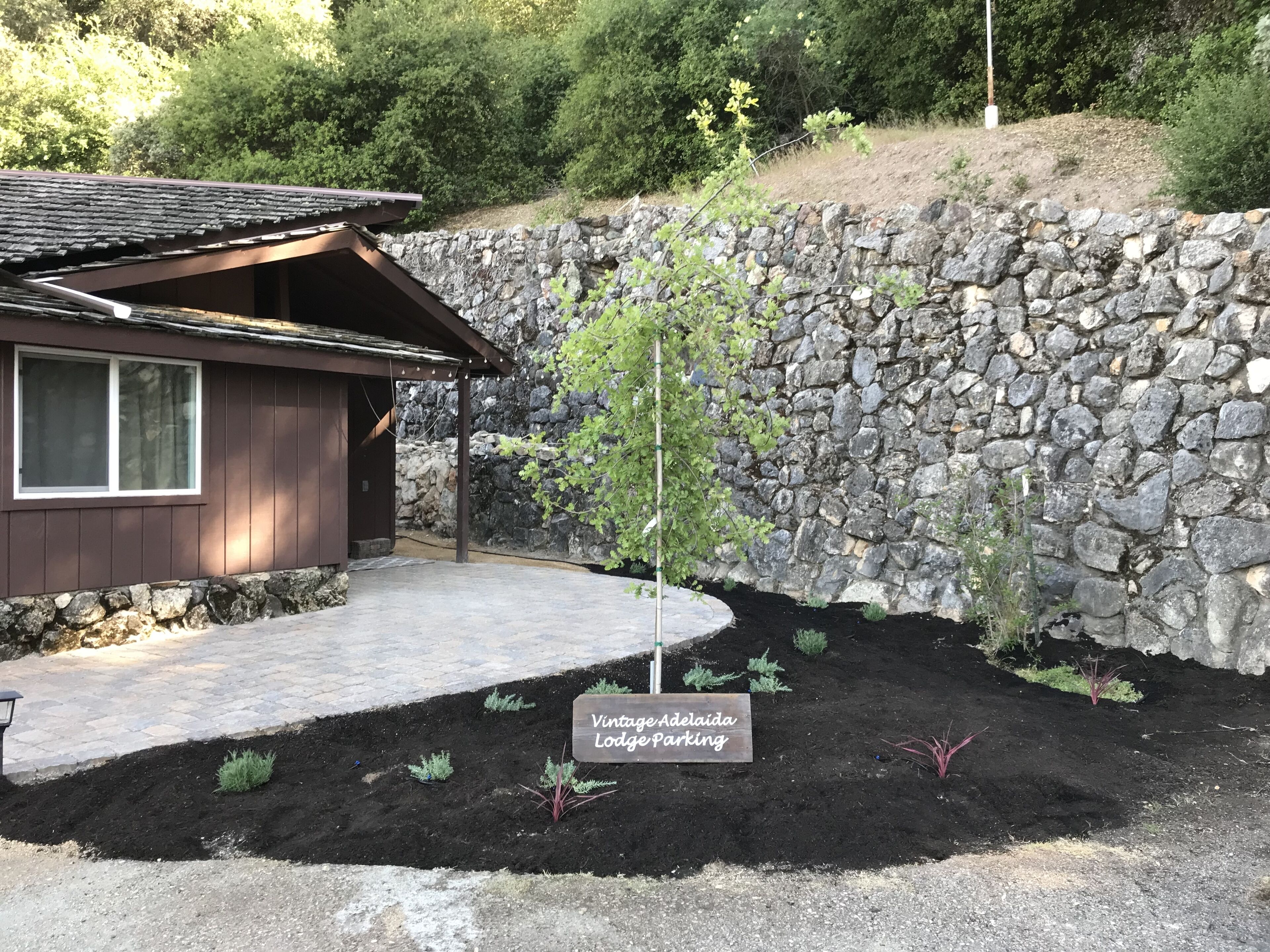 A lodge with a stone wall, a small garden with labeled plants, and a paved driveway.