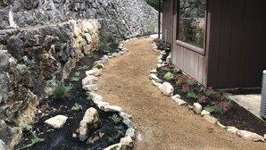 Um caminho que leva a um edifício, com uma parede de pedra de um lado e um jardim com pedras e plantas do outro.
