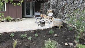 Uma cabana rústica com uma parede de pedra, um alpendre com duas cadeiras e um barril, e uma porta que dá para o interior.