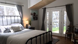 Quarto com cama, mesa de cabeceira, luminária, cadeira e vista através de uma porta de vidro.