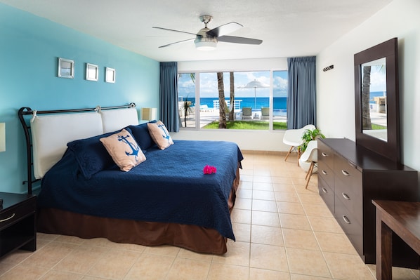 A bedroom with a bed, a ceiling fan, a view of the beach, and a window with curtains.