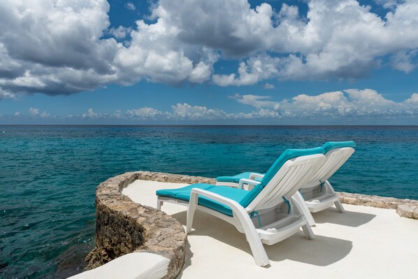 Two white lounge chairs with blue cushions on a stone platform by the sea.