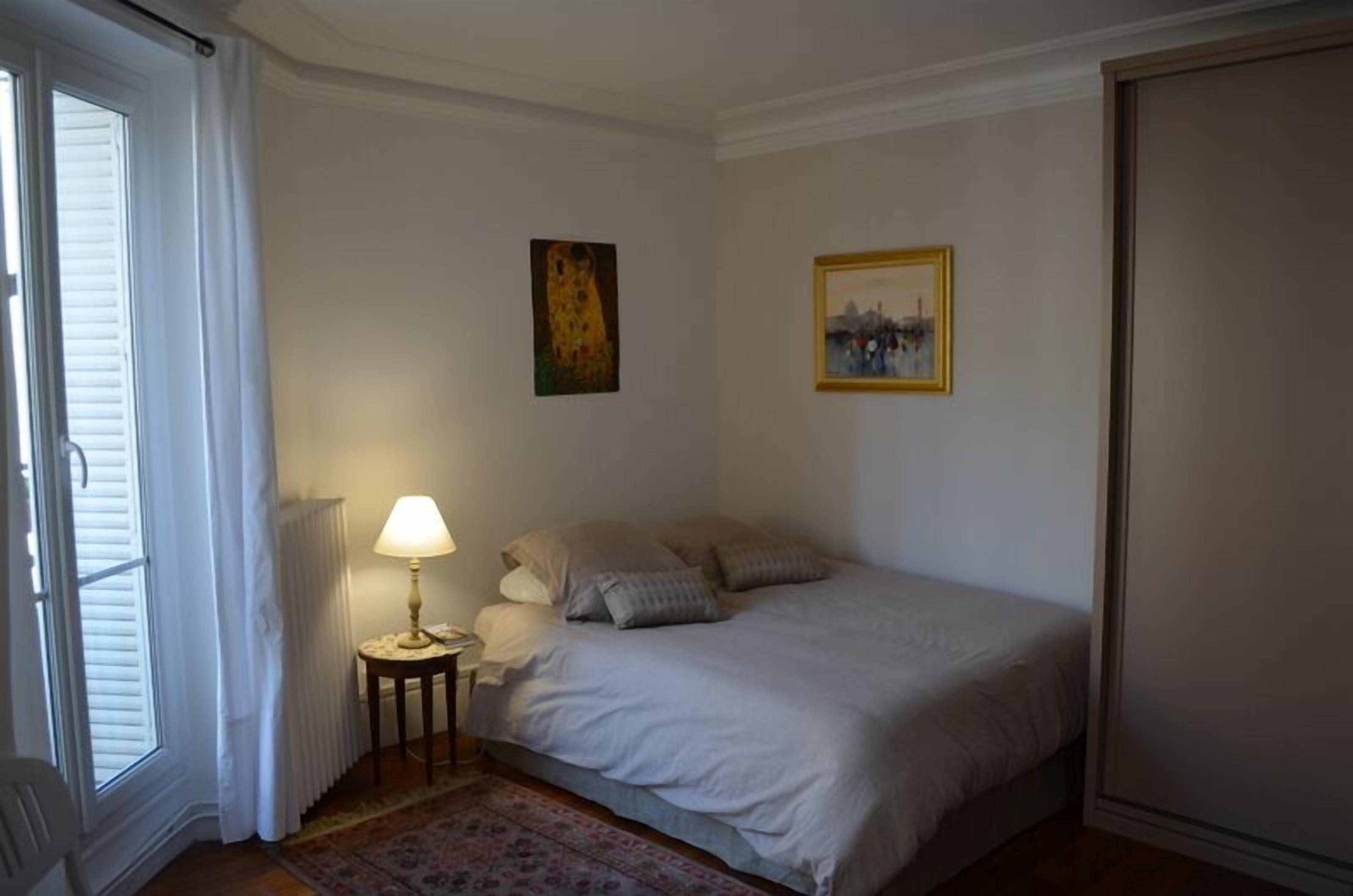 A bedroom with a bed, a nightstand with a lamp, a radiator, and two framed pictures on the wall.