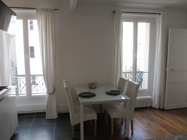 Un coin repas avec une table et des chaises blanches, une fenêtre avec des rideaux et une vue sur l’extérieur.