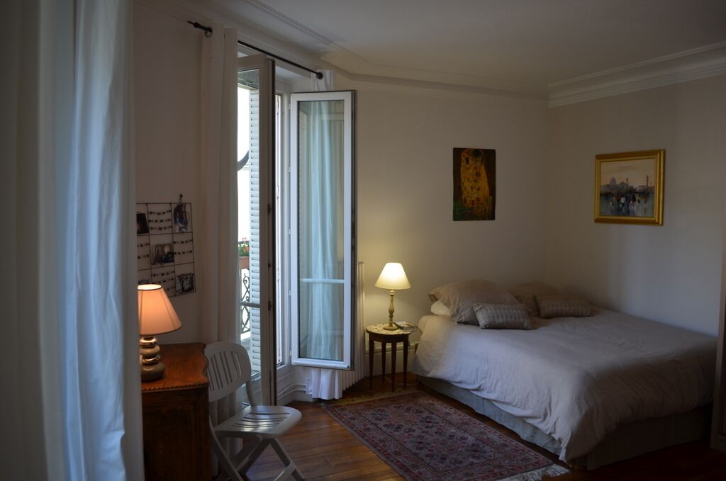 A bedroom with a bed, a desk with a lamp, a chair, a small table with a lamp, and a window with curtains.