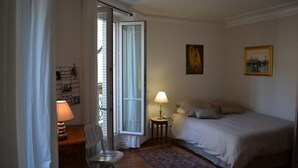 Ett sovrum med en säng, ett skrivbord med en lampa, en stol, ett litet bord med en lampa och ett fönster med gardiner.