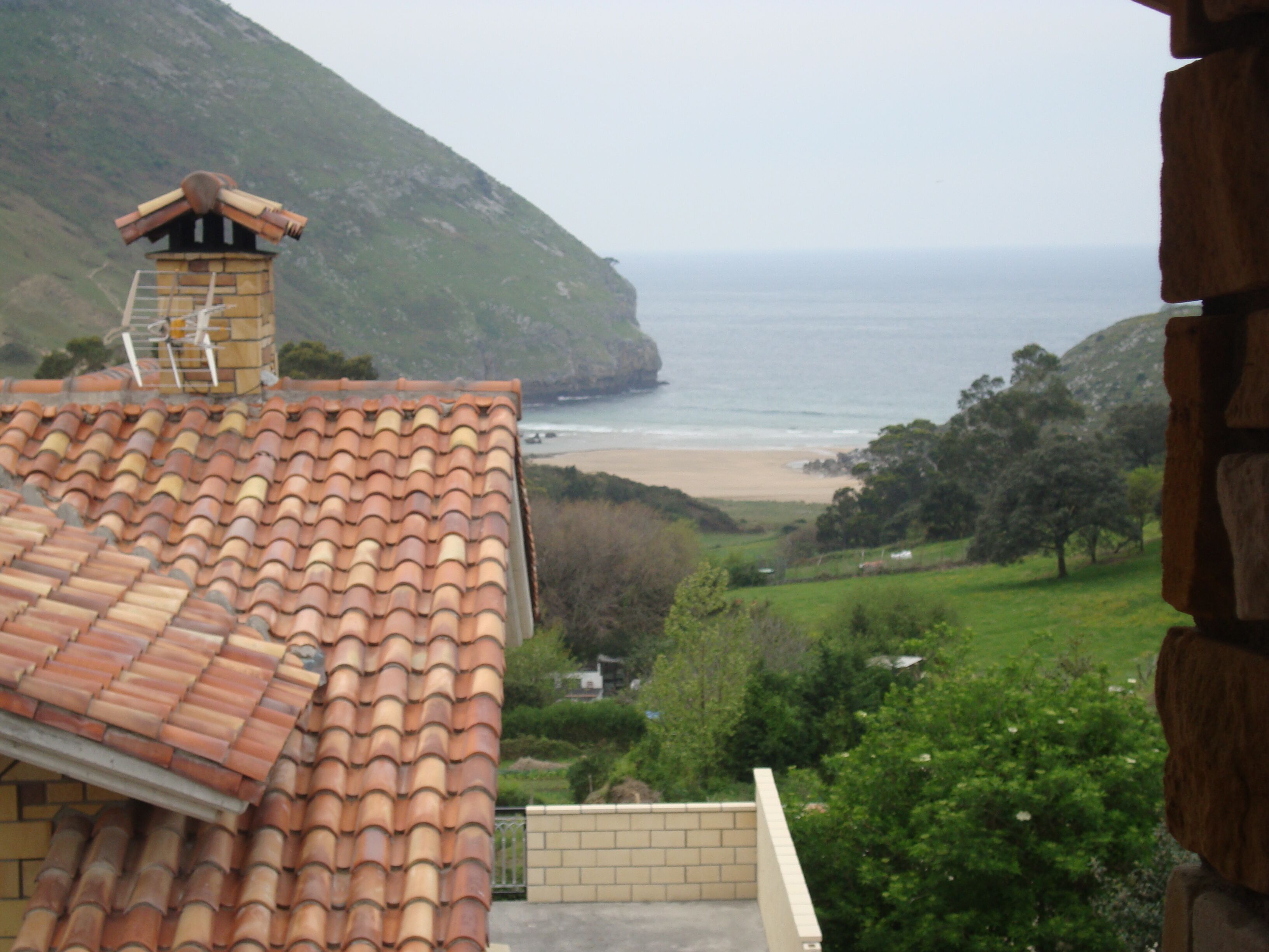 A coastal view with a tiled roof, a chimney, and a beach in the distance.